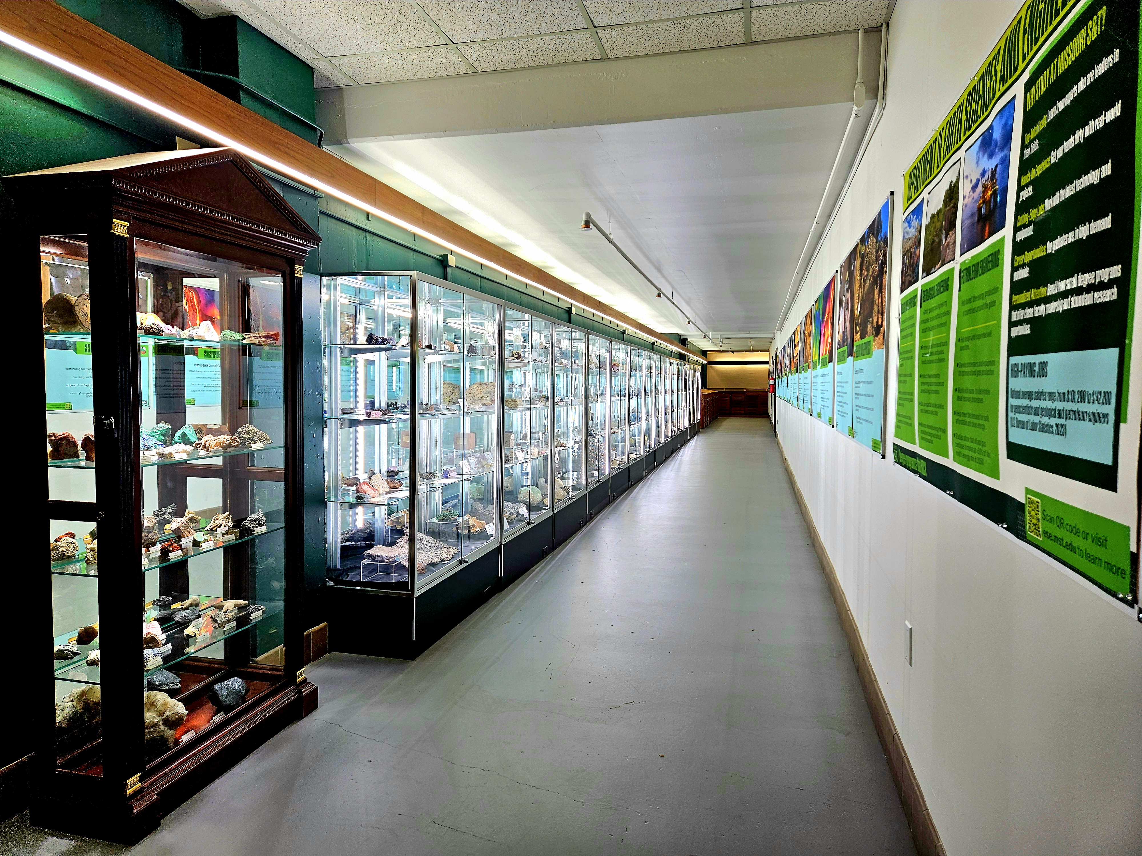 The hallway of the S&T Mineral Museum, with a row of brightly-lit display cases containing minerals against the left wall, and informational posters on the right wall.