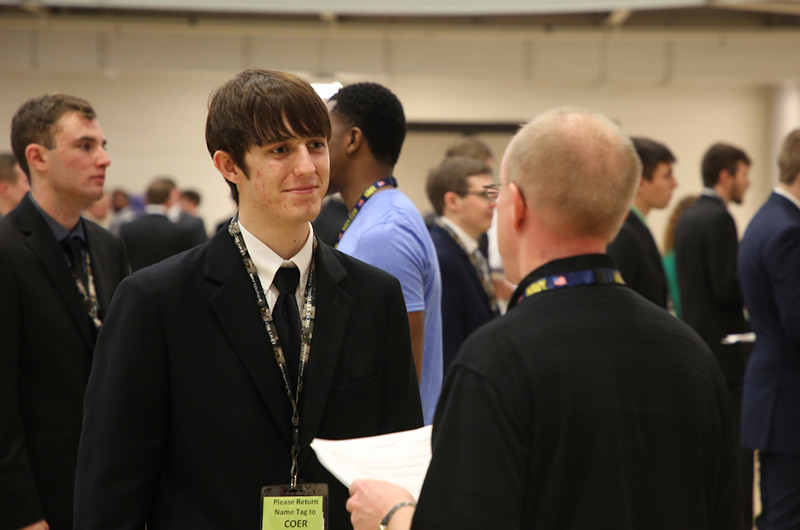 Student in a suit conversing with a professional at a career fair, surrounded by other students and professionals in the background